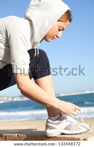 Close up profile portrait of a young sports woman doing up her trainers shoe laces against an intense blue sky by a golden beach.