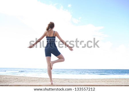 Rear view of a young woman in a yoga tree position on a golden beach with her arms open against a blue sky.
