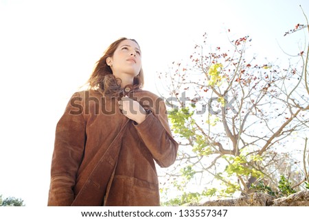 Low perspective portrait of a young attractive woman wearing a leather coat and covering herself during a sunny winter day in a park.