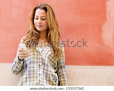 Portrait of an attractive young woman leaning on a colorful old city wall using a high tech smartphone and wearing a checked shirt, smiling.