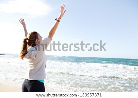 Side rear view of a young sporty woman stretching her arms up while standing on a beach by the shore and against a blue sky.