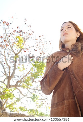 Low perspective portrait of a young attractive woman wearing a leather coat and covering herself during a sunny winter day in a park.
