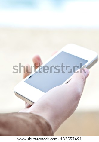 Close up detail view of a woman\'s hand holding a modern smart phone with a blank screen against a neutral background and the sea.