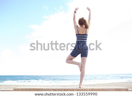 Rear view of a young woman in a yoga tree position on a golden beach with her arms open against a blue sky.