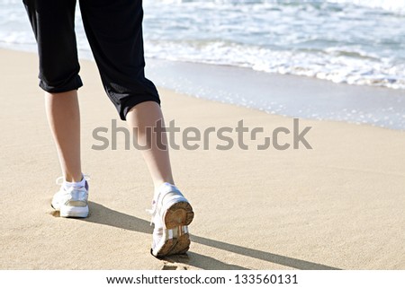 Graphic low section view of a young woman\'s legs and feet running along the sea shore exercising, wearing sport trainers. Rear view.