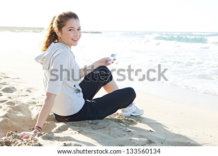 Portrait of a young woman sitting on a white sand beach shore, holding a music player and listening to music with her head phones, turning and smiling at the camera.