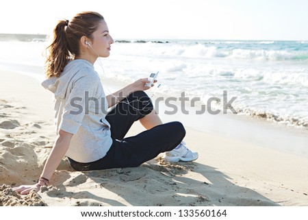 Profile portrait view of a young woman sitting on a fine sand beach shore, holding a music player and listening to music with her head phones being thoughtful.