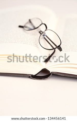 Detail view of a pair of reading glasses with a black frame laying on the pages of an open hard back book on a white desk.