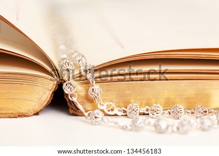 Close up profile detail view of an open Bible book with a silver beads rosary laying between the pages on a white desk.