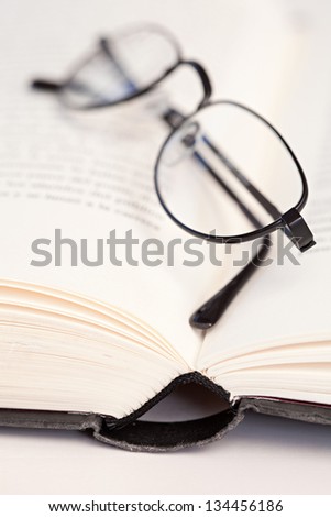 Close up view of a pair of reading glasses with a black frame laying on the pages of an open hard back book on a white desk.