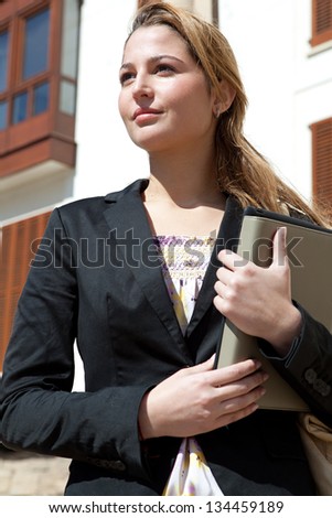 Close up portrait of a successful businesswoman holding a digital tablet pad and a folder while proudly standing near a classic city office buildings.
