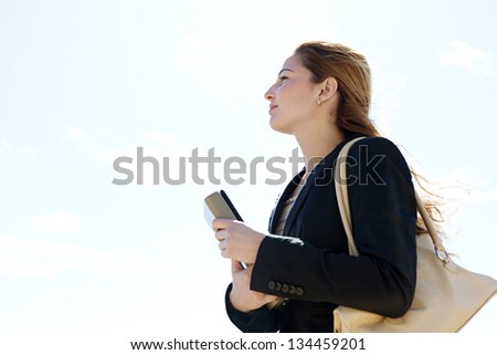 Profile portrait view of a young successful businesswoman carrying work folders and wearing a black suit, looking ahead against a blue sky background.