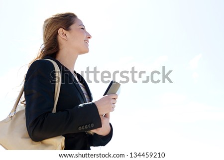 Profile portrait view of a young successful businesswoman carrying work folders and wearing a black suit, looking ahead against a blue sky, smiling.