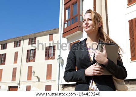 Portrait of a successful businesswoman holding a digital tablet pad and a folder while proudly standing near classic city buildings.