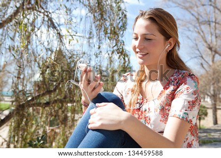 Close up portrait of a young attractive woman using a smartphone to listen to music while sitting in a park with a blue sky during a sunny day.