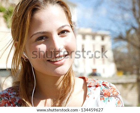 Close up portrait of a young woman using a smartphone to listen to music with her head phones, sitting outdoors in the city on a sunny day.
