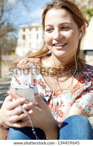 Close up portrait of a young woman using a smartphone to listen to music with her head phones, sitting outdoors in the city on a sunny day.