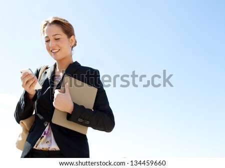 Portrait Of An Attractive Young Professional Woman Using A Smartphone While Standing Against A Sunny Blue Sky, Smiling.