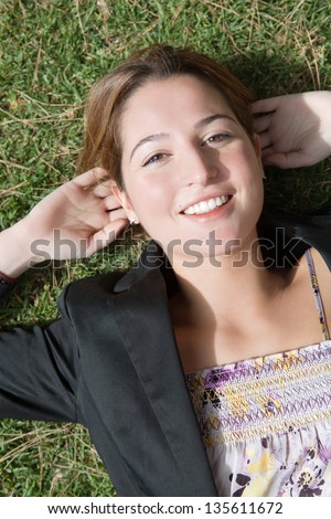 Over head view of a young professional woman laying down on green grass in a park with her hands behind her head, smiling at the camera.