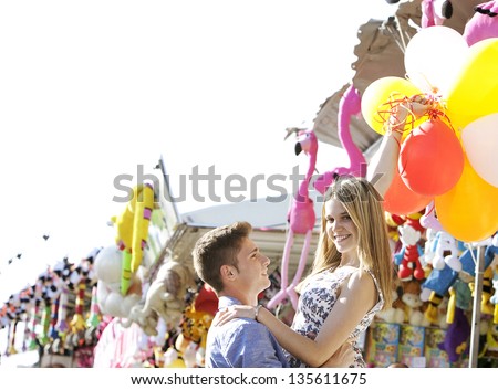 Teenage boy lifting teenage girl up while she\'s holding colorful balloons up in the air in an amusement park arcade with games and toys.