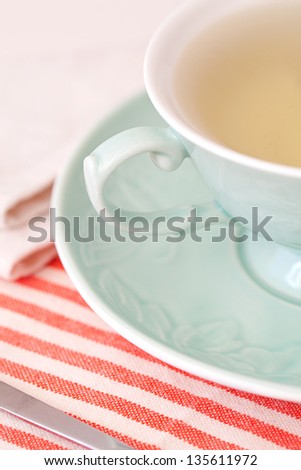 Close up view of a cup of hot tea in a delicate green porcelain cup on a stripy red and white table cloth in a home kitchen.