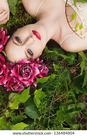 Over head view of a beautiful young woman laying down on a bed of green lush leaves, wearing a large blossom flower had during spring.
