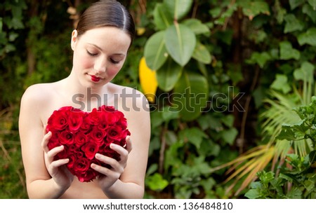 Attractive young woman nude in a green lush garden holding a red roses heart in front of her chest, wearing red lipstick and being thoughtful.