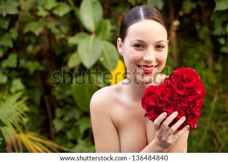 Attractive young woman nude in a green lush garden holding a red roses heart in front of her chest, wearing red lipstick and smiling.