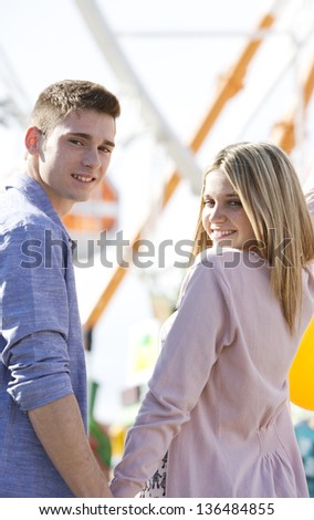 Portrait of an attractive teenage couple in an amusement park with rides, turning around to look at the camera while holding hands, smiling.