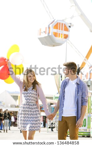 Young joyful couple having fun in a fun fair ground, smiling and holding colorful balloons.