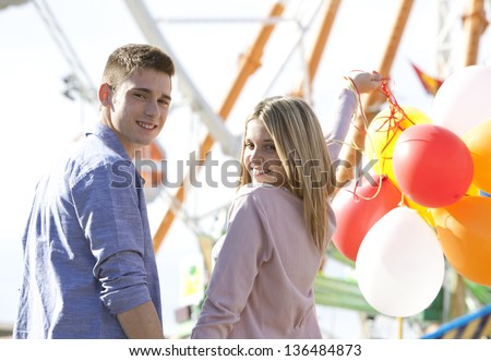 Attractive teenage couple in an amusement park with rides, holding balloons up in the air and turning around to look at the camera smiling.