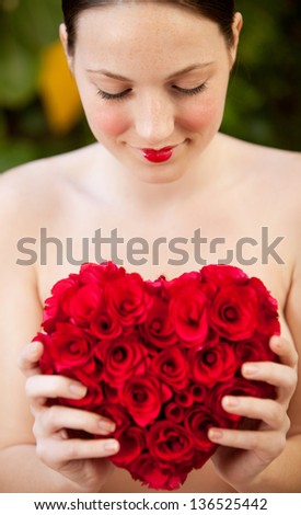Attractive young woman naked in a green lush garden holding a red roses heart in front of her chest, wearing red lipstick, smiling.