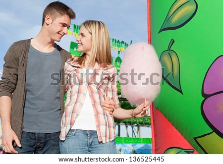 Young couple walking holding each other in an attractions park arcade with cotton candy floss and rides in the background.