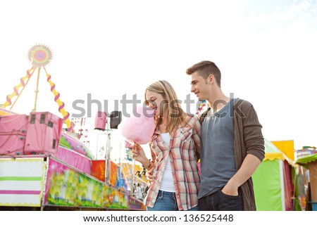 Portrait of a young teenage couple walking and eating cotton candy floss while enjoying visiting a funfair ground with rides in the background, smiling.