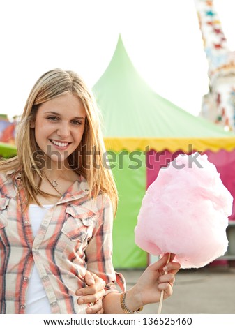 Portrait of a young teenage girl walking and eating cotton candy floss while enjoying visiting a funfair ground with rides in the background, smiling.