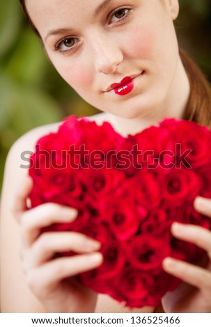 Close up view of an attractive young woman nude in a green lush garden holding a red roses heart, wearing red lipstick and being thoughtful.