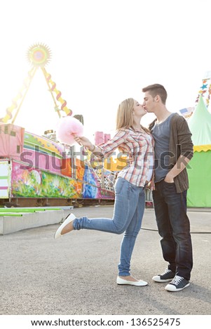 Young fun couple kissing and holding cotton candy floss sweet while visiting an amusement park during a sunny day.