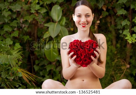 Attractive young woman sitting naked in a green lush garden holding a red roses heart in front of her chest, wearing red lipstick, smiling.