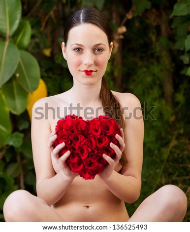 Attractive young woman sitting naked in a green lush garden holding a red roses heart in front of her chest, wearing red lipstick.