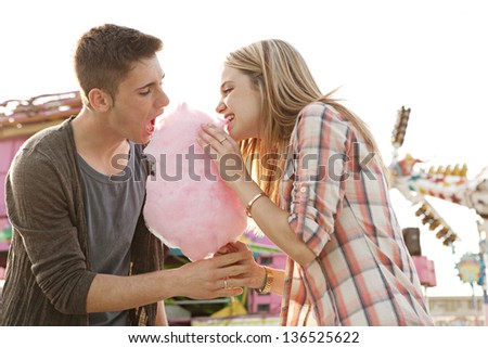 Young fun couple biting into a cotton candy floss sweet at the same time while visiting an amusement park during a sunny day.