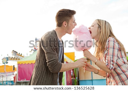Young fun couple biting into a cotton candy floss sweet at the same time while visiting a colorful amusement park during a sunny day.