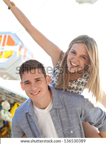Joyful young couple being playful at an amusement park, with young man carrying his girlfriend on his back near a ferris wheel.