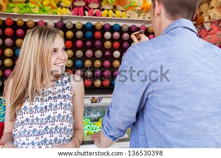 Young fun couple playing darts games in an amusement park arcade during a sunny day, with toys and prices around them, smiling.