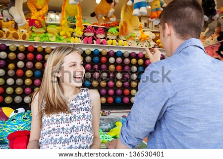 Young fun couple playing darts games in an amusement park arcade during a sunny day, with toys and prices around them, smiling.