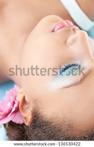 Close up over head view of a young teenage girl wearing blue make up and a pink flower in her head while laying down and being thoughtful.