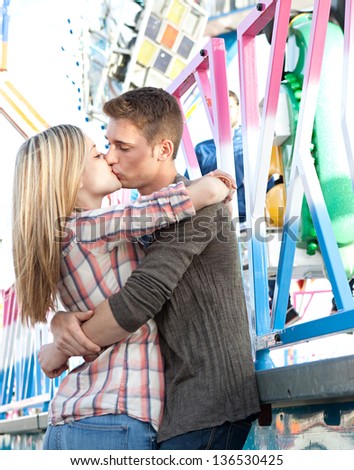 Young attractive couple hugging and kissing while visiting an amusement park arcade during a sunny day, standing by a colorful ride.
