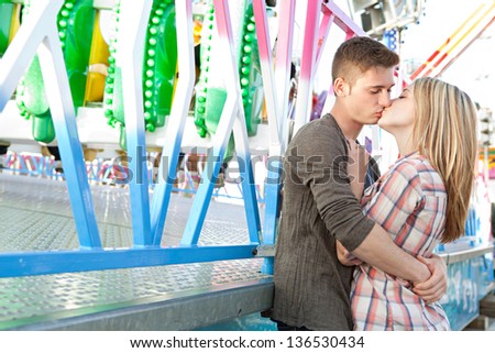 Young attractive couple hugging and kissing while visiting an amusement park arcade during a sunny day, standing by a colorful ride.
