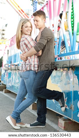 Young attractive couple hugging and smiling while visiting a colorful amusement park arcade during a sunny day.
