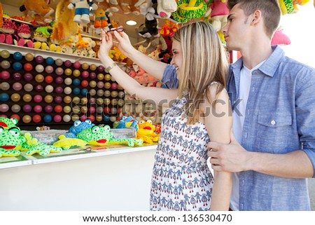 Teenage boy helping his girlfriend aim while playing a darts game at an amusement park arcade with prices and toys around them. Side view.