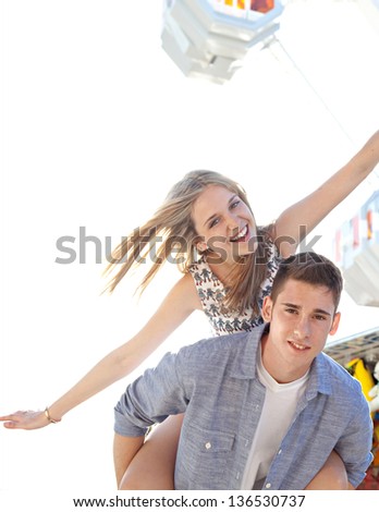 Joyful young couple being playful at an amusement park, with young man carrying his girlfriend on his back near a ferris wheel.